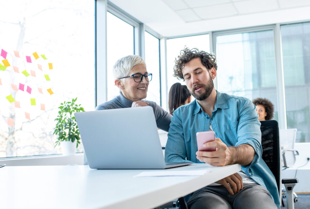 A relaxed office atmosphere, work break. A man is holding a phone and talking to a senior colleague. There are two more women behind and there are sticky notes on a large window.